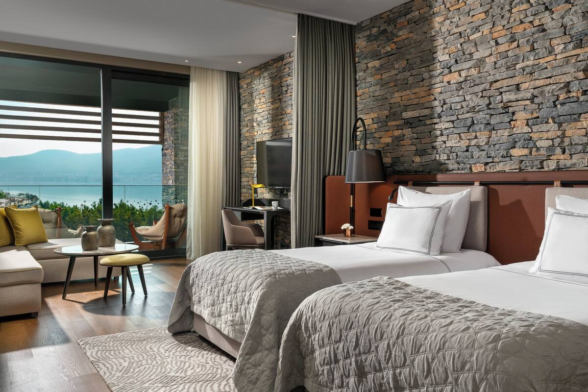 A hotel room with two beds, a desk, a chair, and a view of mountains.
