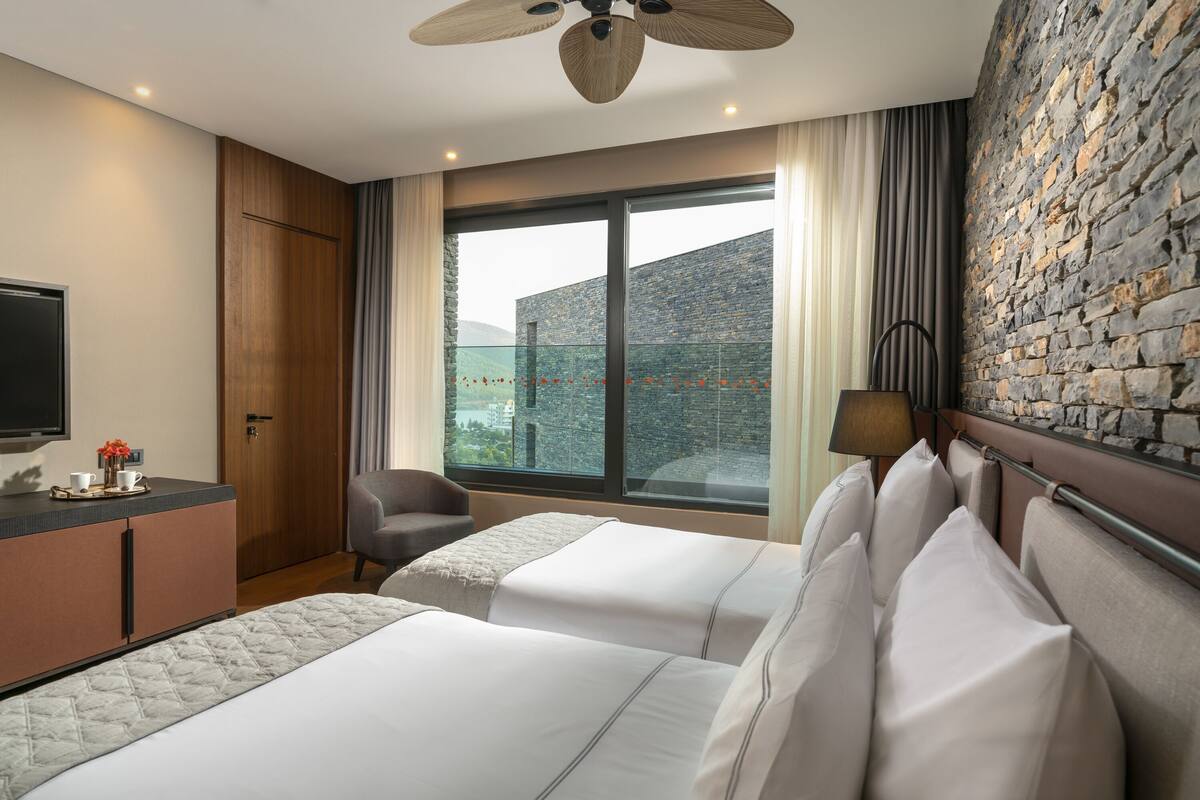 A hotel room with two beds, a desk, a chair, a TV, and a large window with a view.