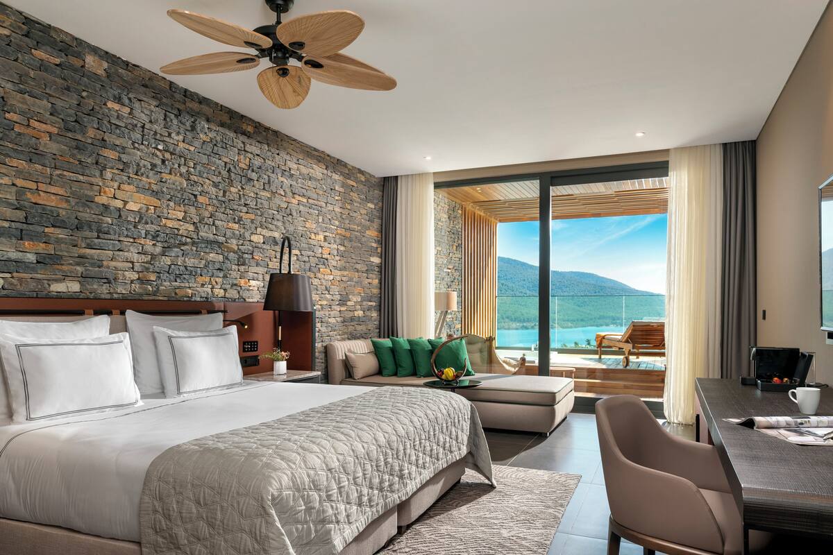 A modern hotel room with a stone wall, a large bed, a seating area with a view of the mountains, and a balcony with a table and chairs.