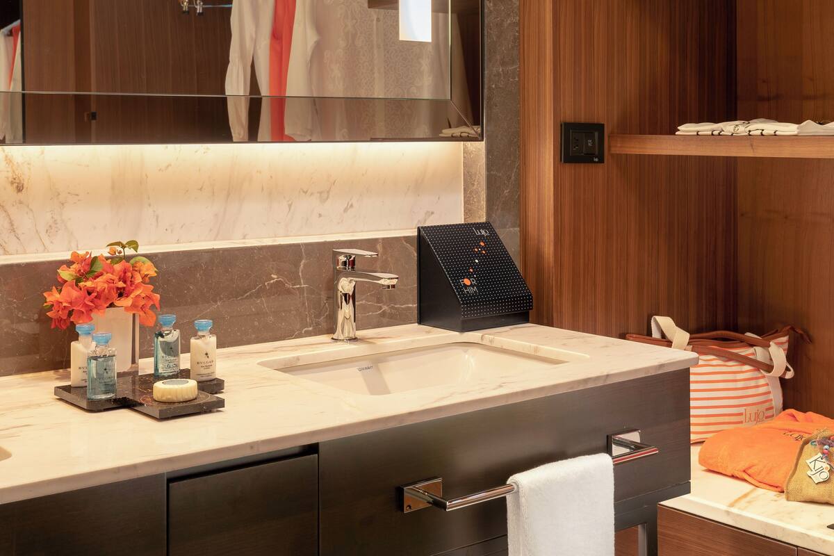 A modern bathroom with a large mirror, a sink, and a vanity. There are toiletries on the counter, a towel rack, and a shelf with a bag and a bottle.