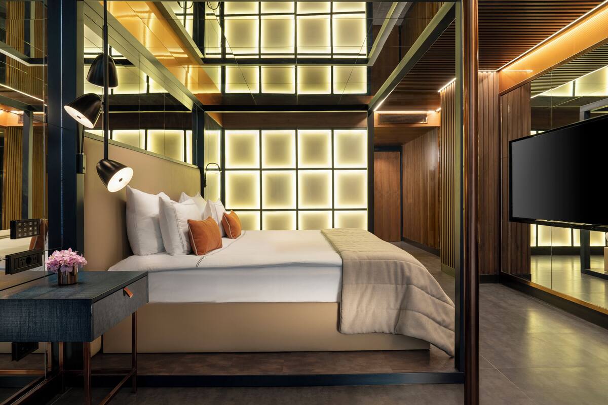 A modern hotel room with a large bed, a desk, a TV, and a unique ceiling design.