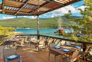 Eine Holzterrasse mit Tischen und Stühlen, die auf ein Gewässer mit Booten und eine Bergkulisse blickt.