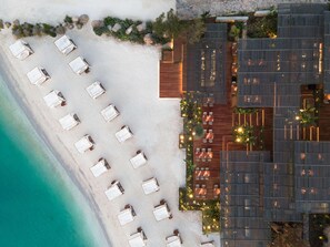 Ein Luftbild eines Strandhotels mit Sandstrand, Reihen aus weißen Liegestühlen und einem modernen Gebäude mit Holzdetails und viel Grün.