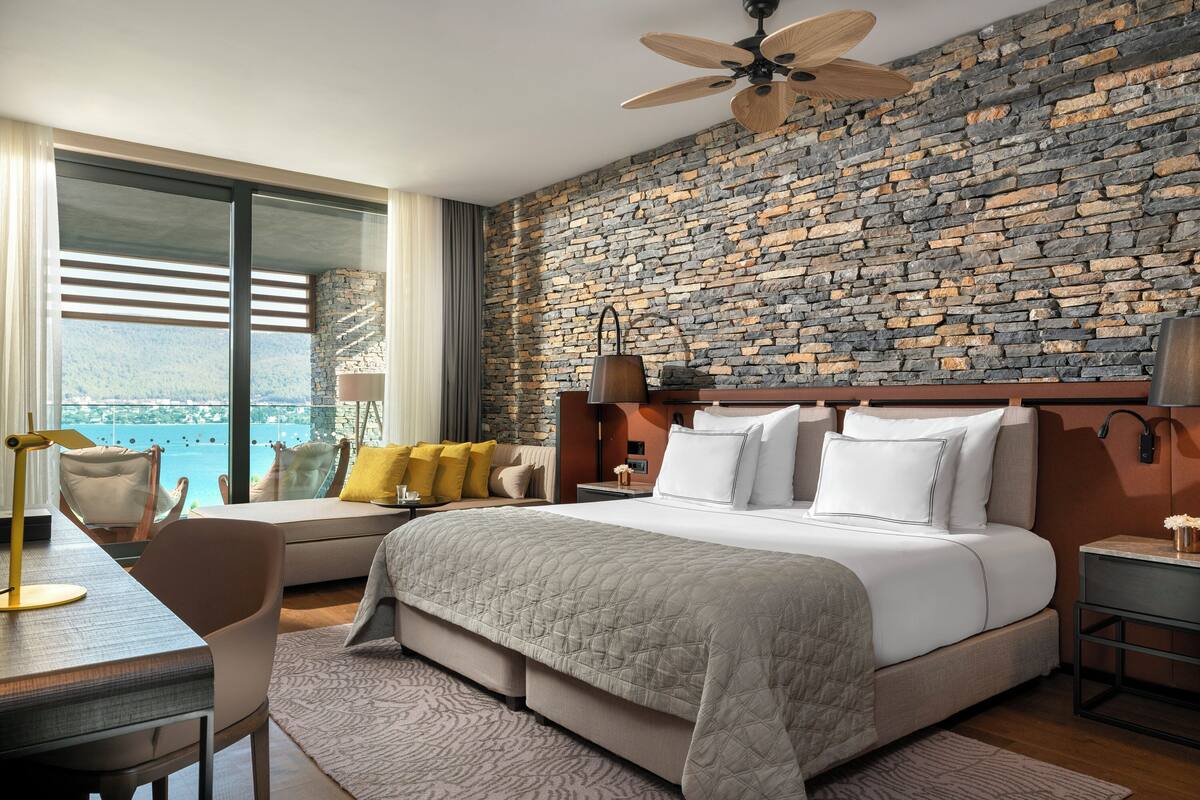 A modern hotel room with a large bed, a desk, and a view of the sea.
