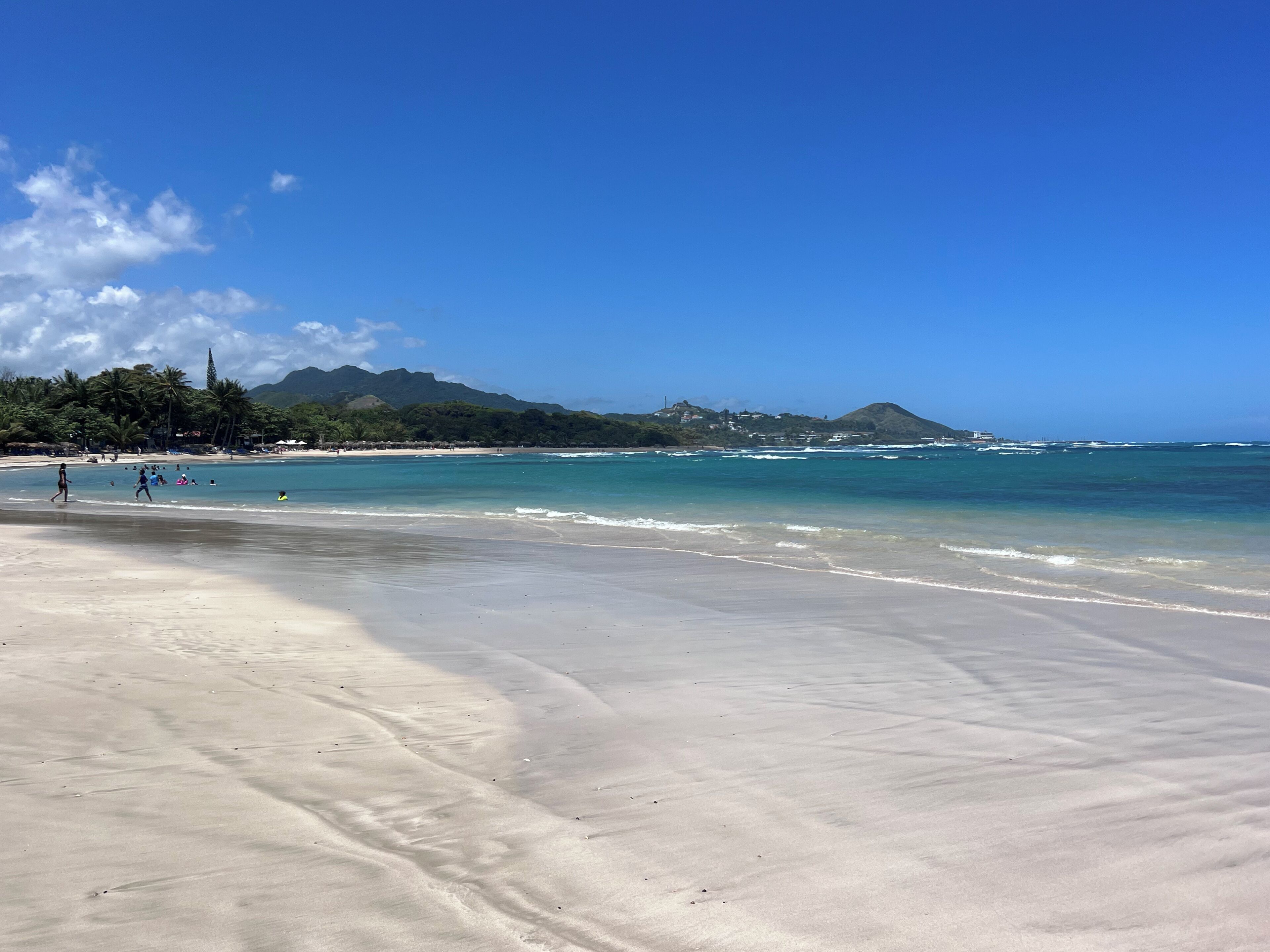 Ein Strand mit klarem Wasser, sandigem Ufer und einem Berg im Hintergrund.