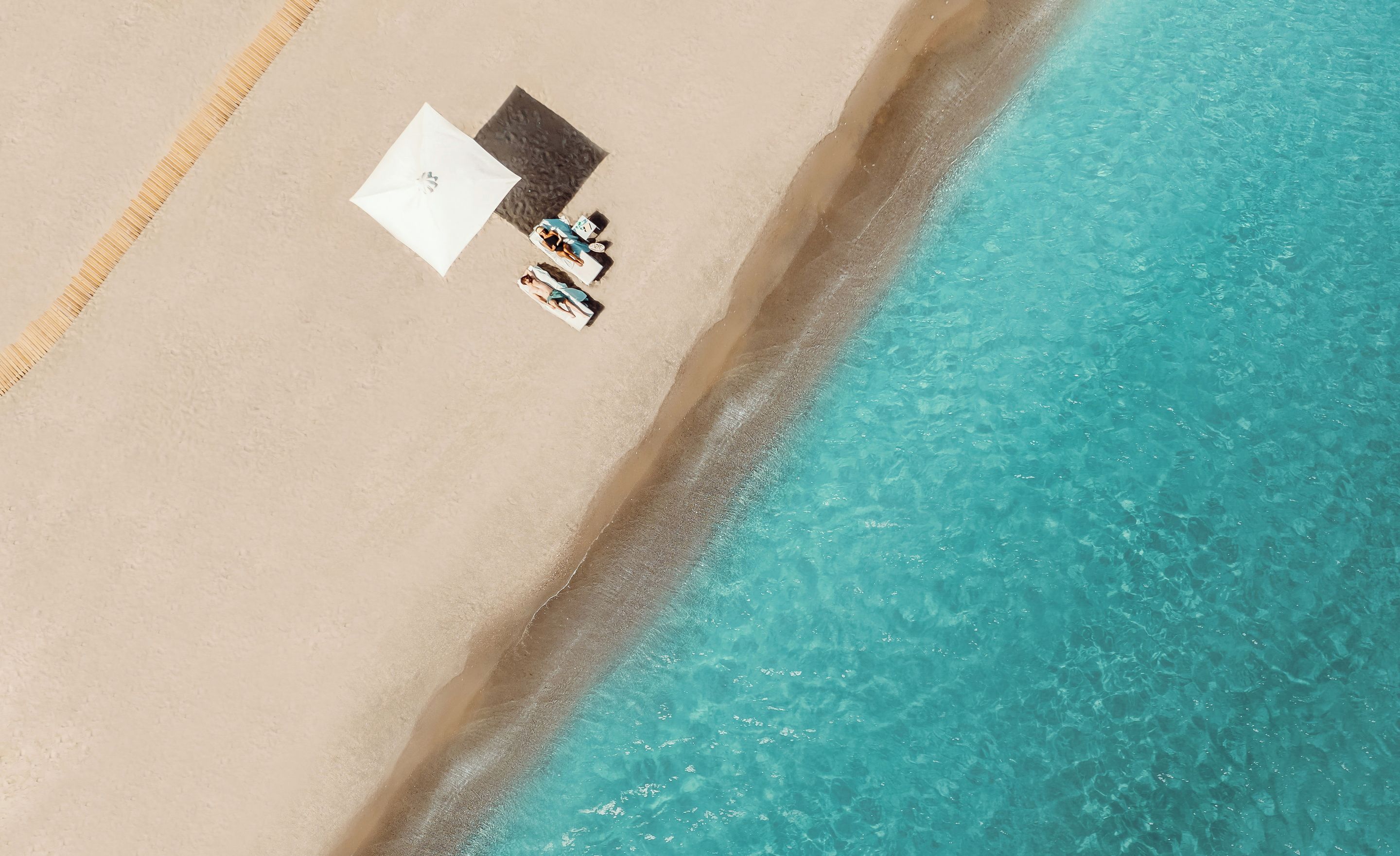 An aerial view of a beach with a white umbrella and a pair of shoes on the sand, and a clear blue ocean beside it.