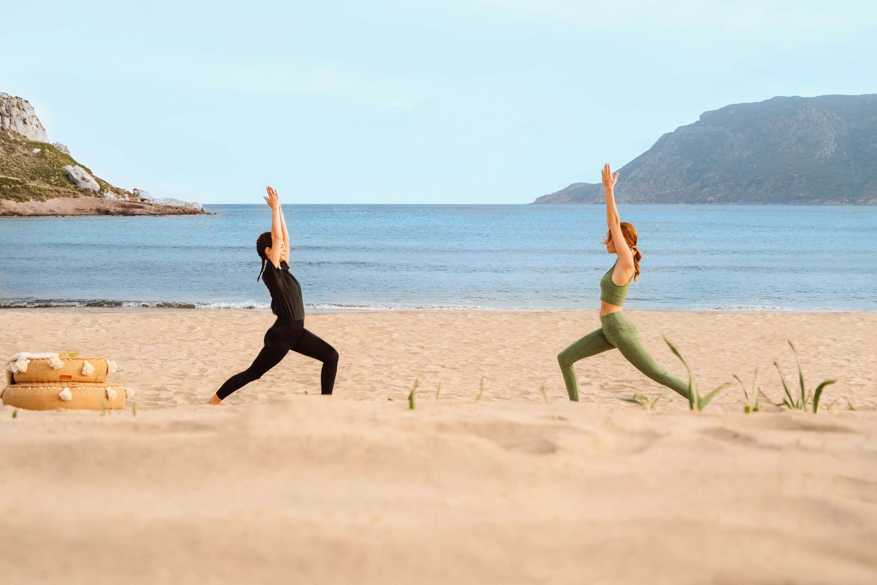 Twee personen beoefenen yoga op een strand, met een helder uitzicht op de zee en heuvels in de verte.