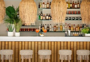 Una zona de bar con lámparas de fibra tejida, estantes llenos de botellas de licor y taburetes de macramé.