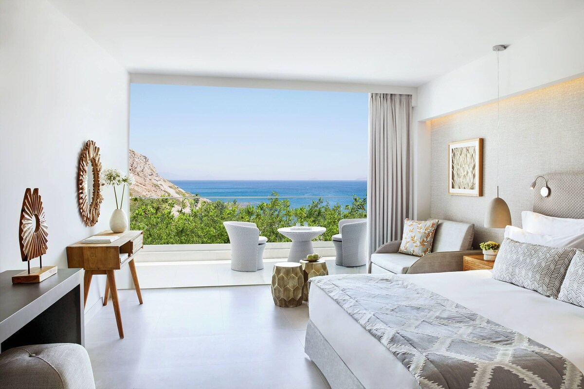 A modern bedroom with a large window, a bed, a desk, and a seating area with a view of the ocean.