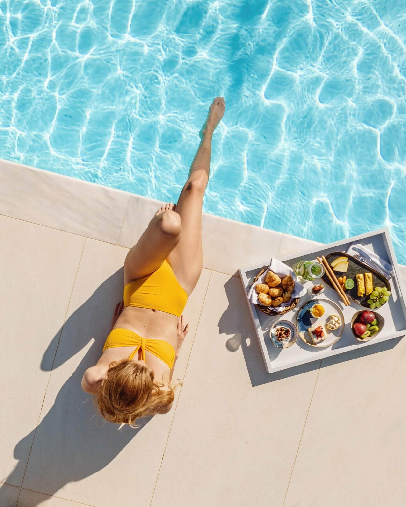 Una persona recostada en una silla de salón junto a la piscina, con una bandeja de comida.