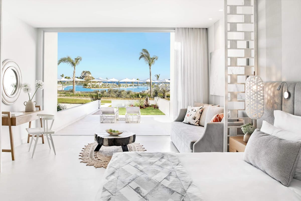 Una habitación de hotel moderna con balcón, una cama, un sofá, un escritorio y vistas a una piscina y palmeras.