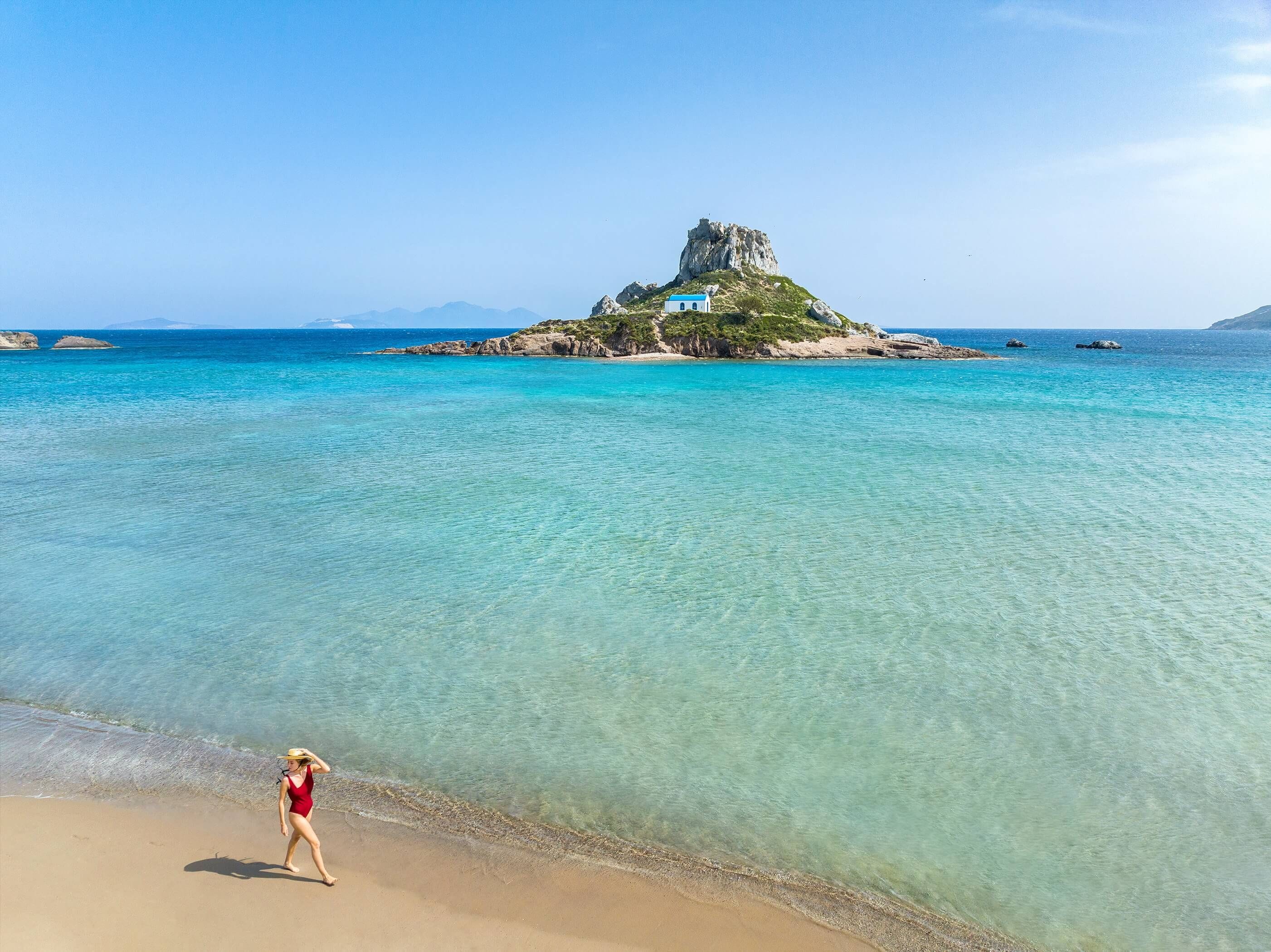 A beach with clear turquoise water, a rocky island with a building, and a person walking on the shore.