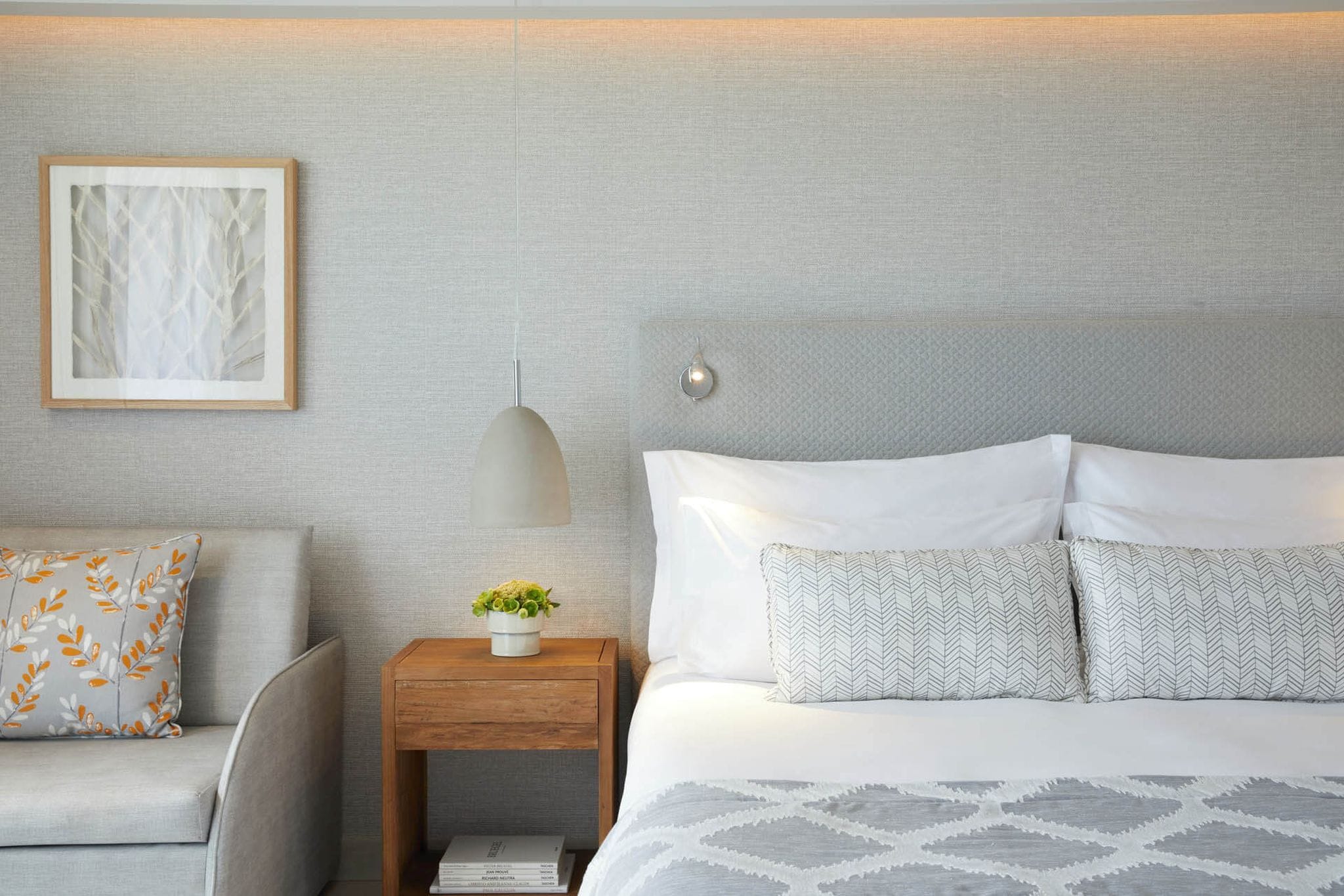 A neatly arranged bedroom with a bed, a nightstand with a lamp, a framed picture on the wall, and a sofa with patterned cushions.
