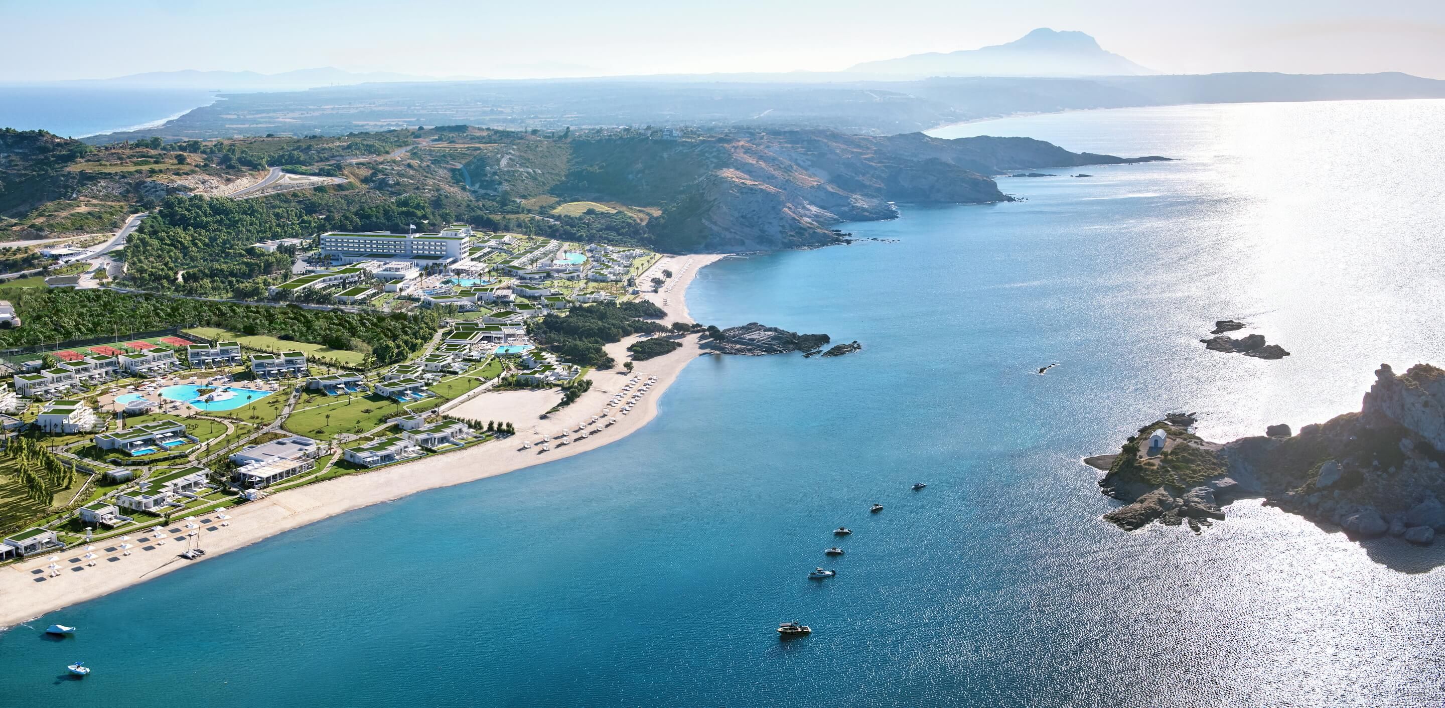 An aerial view of a coastal resort with a beach, swimming pools, and a mountainous backdrop.