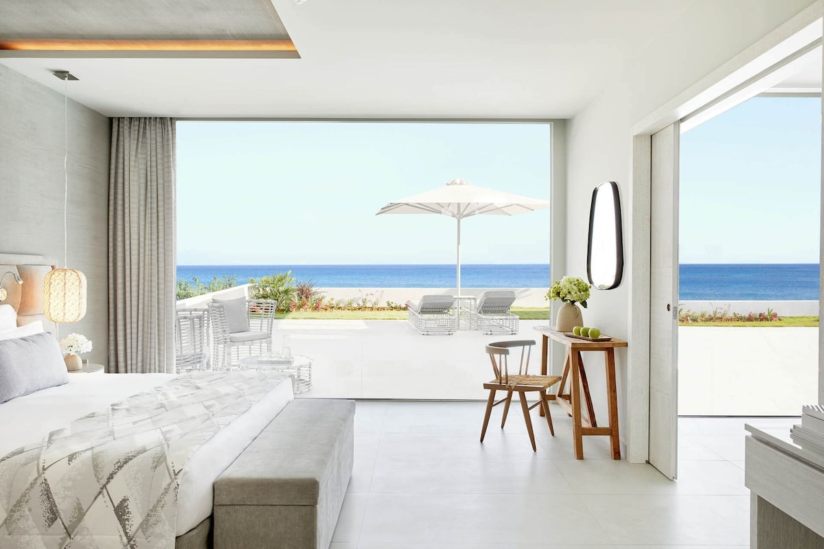 A modern bedroom with a large bed, a balcony with a view of the sea, and a small table with chairs.