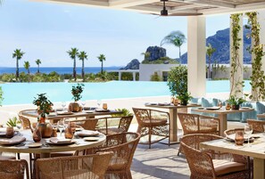 An outdoor dining area with wicker furniture, tables set for dining, and a view of the ocean and palm trees. - Ikos Aria (Kos)