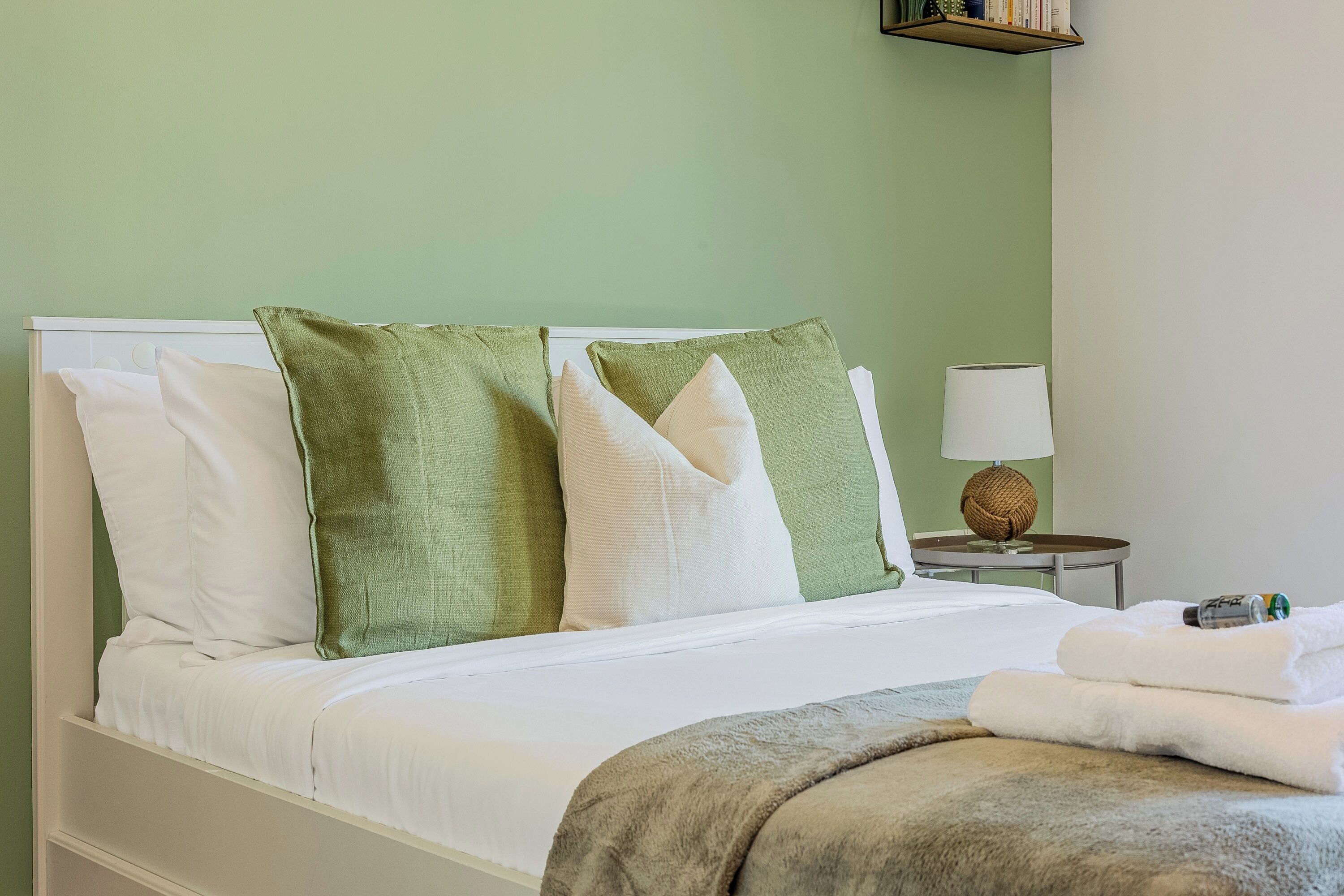 A bed with white and green pillows, a white blanket, and a grey throw.