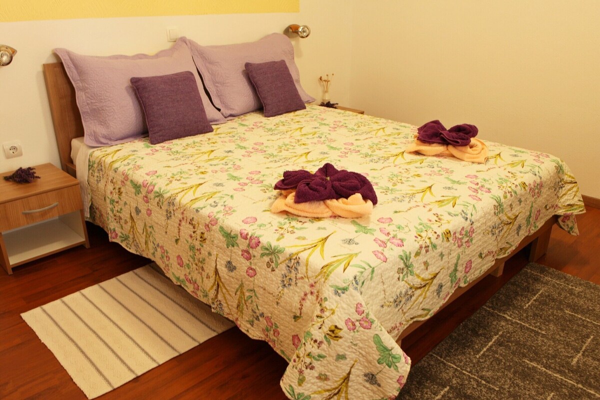 Ein ordentlich gemachtes Bett mit Blumenmuster auf der Bettwäsche, zwei violette Kissen und zwei Handtücher.