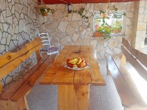 Eine Steinsäule, Holzbank, ein Tisch mit Obst und ein Fenster mit Pflanzen.