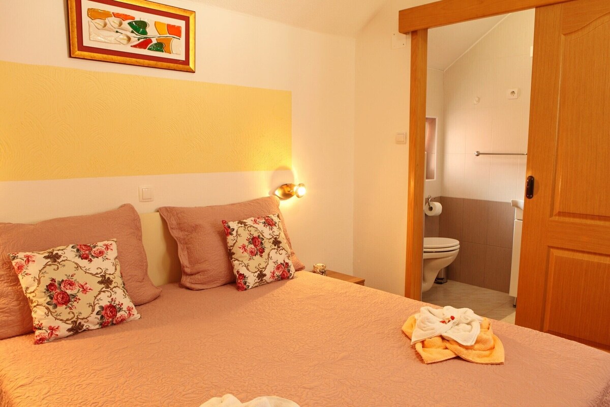 Ein Hotelzimmer mit einem Bett, zwei Kissen, einer Wandleuchte, einem durch eine offene Tür einsehbaren Badezimmer und einem gerahmten Bild an der Wand.