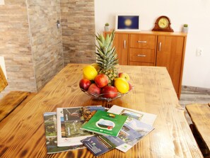 Ein Holztisch mit einer Obstschale, die Äpfel, eine Ananas und eine Zitrone enthält. Auf dem Tisch liegen Broschüren und eine grüne Karte.