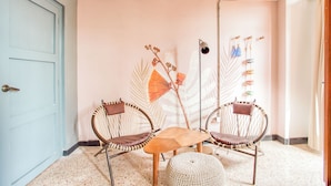 Una habitación con una puerta azul, dos sillas de mimbre, una pequeña mesa de madera, un otoman tejido y una pared con diseño de hojas.