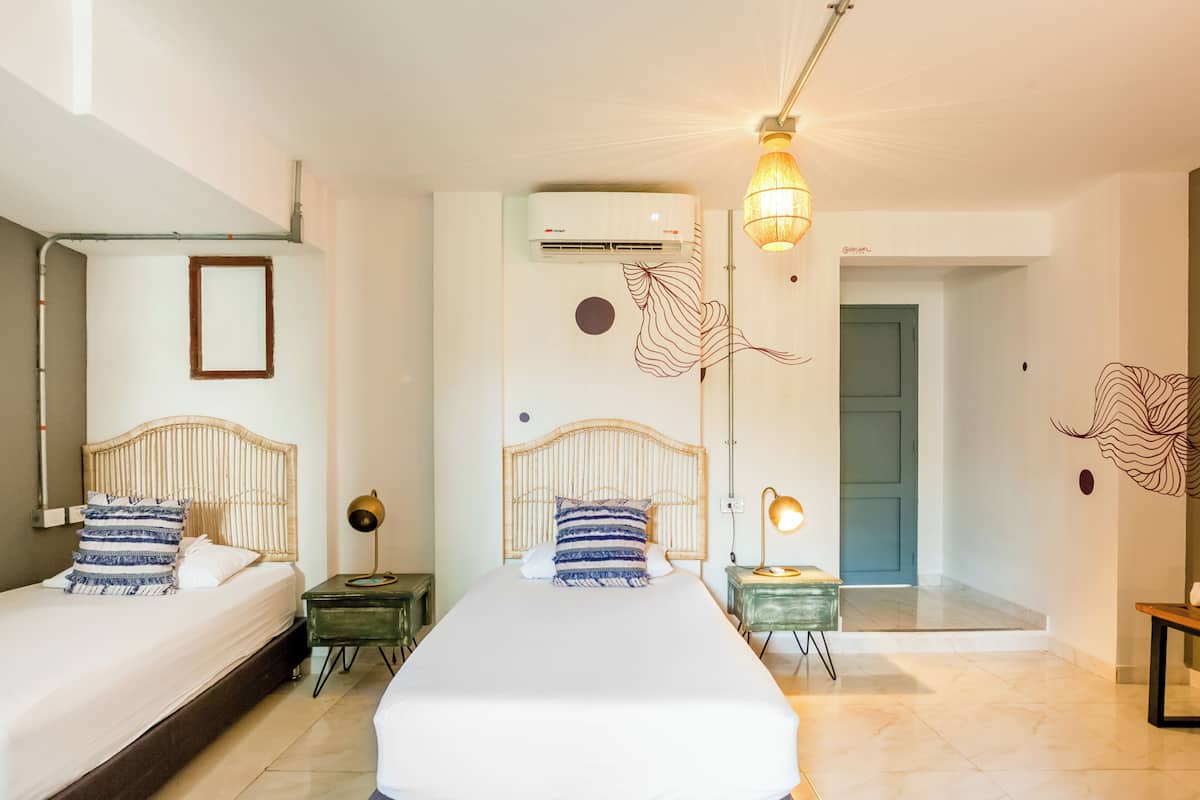 A hotel room with two beds, a mirror, an air conditioning unit, a lamp, and a door leading to another room.