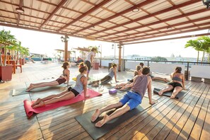 Eine Gruppe von Menschen praktiziert Yoga auf Matten in einem überdachten Freilaufbereich mit Holzboden und Blumentöpfen.