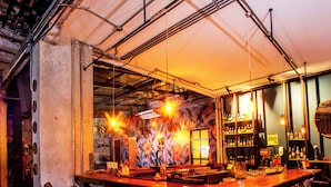 Un bar de diseño rústico, con cañerías a la vista, una barra de madera y sillas de metal.
