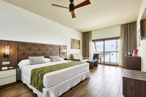 Quarto de hotel com uma cama grande, uma escrivaninha, uma cadeira, um ventilador de teto e vista para o mar pelas janelas.