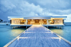 Un hotel moderno con una terraza de madera que conduce a la entrada, estructura sobre el agua y vista despejada del océano.
