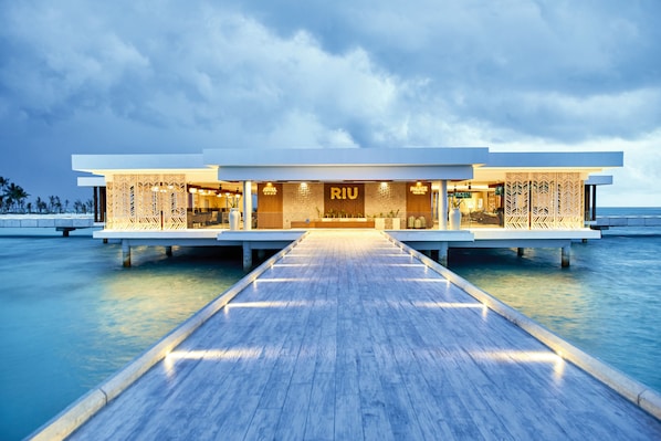Un hotel moderno con una terraza de madera que conduce a la entrada, estructura sobre el agua y vista despejada del océano.