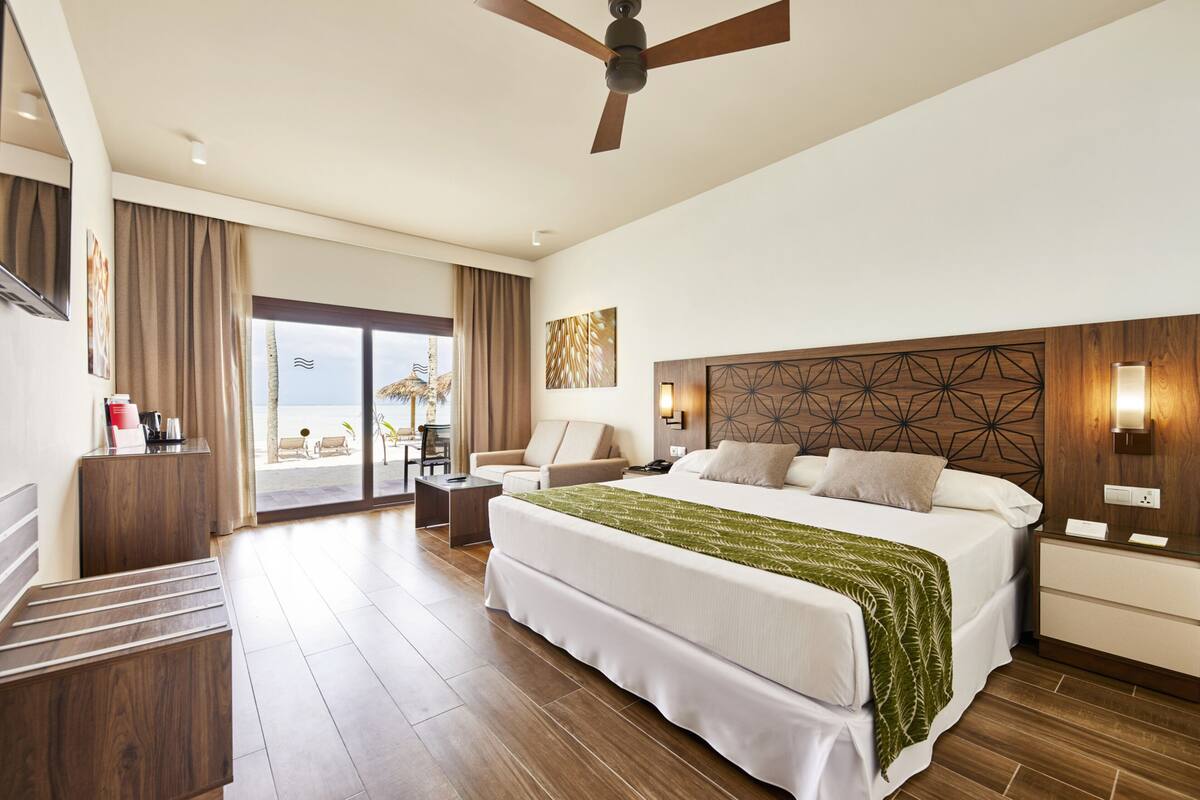 A hotel room with a large bed, a ceiling fan, a desk, a chair, and a view of the beach through the glass door.