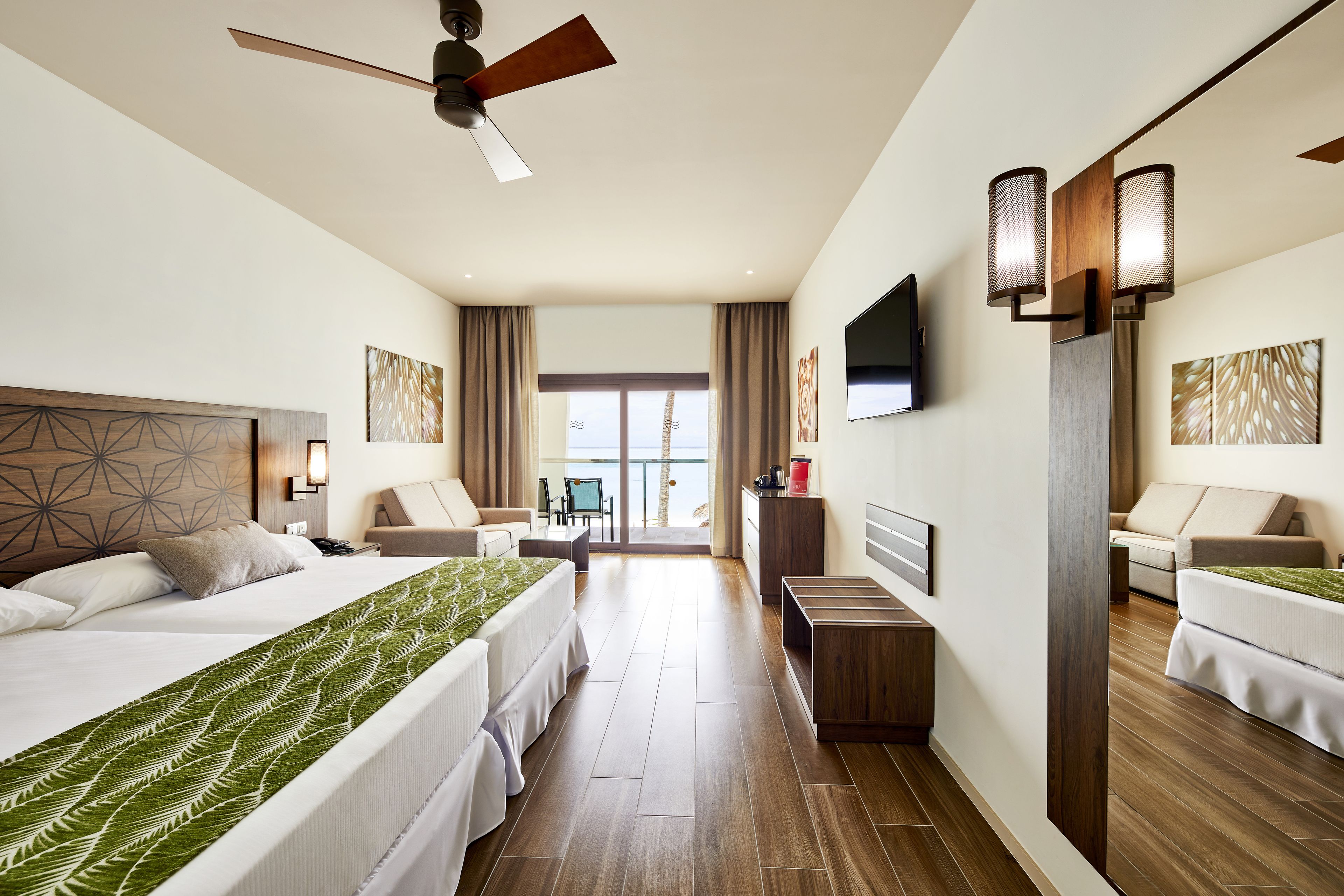 A hotel room with two beds, a desk, a chair, a TV, and a view of the sea.