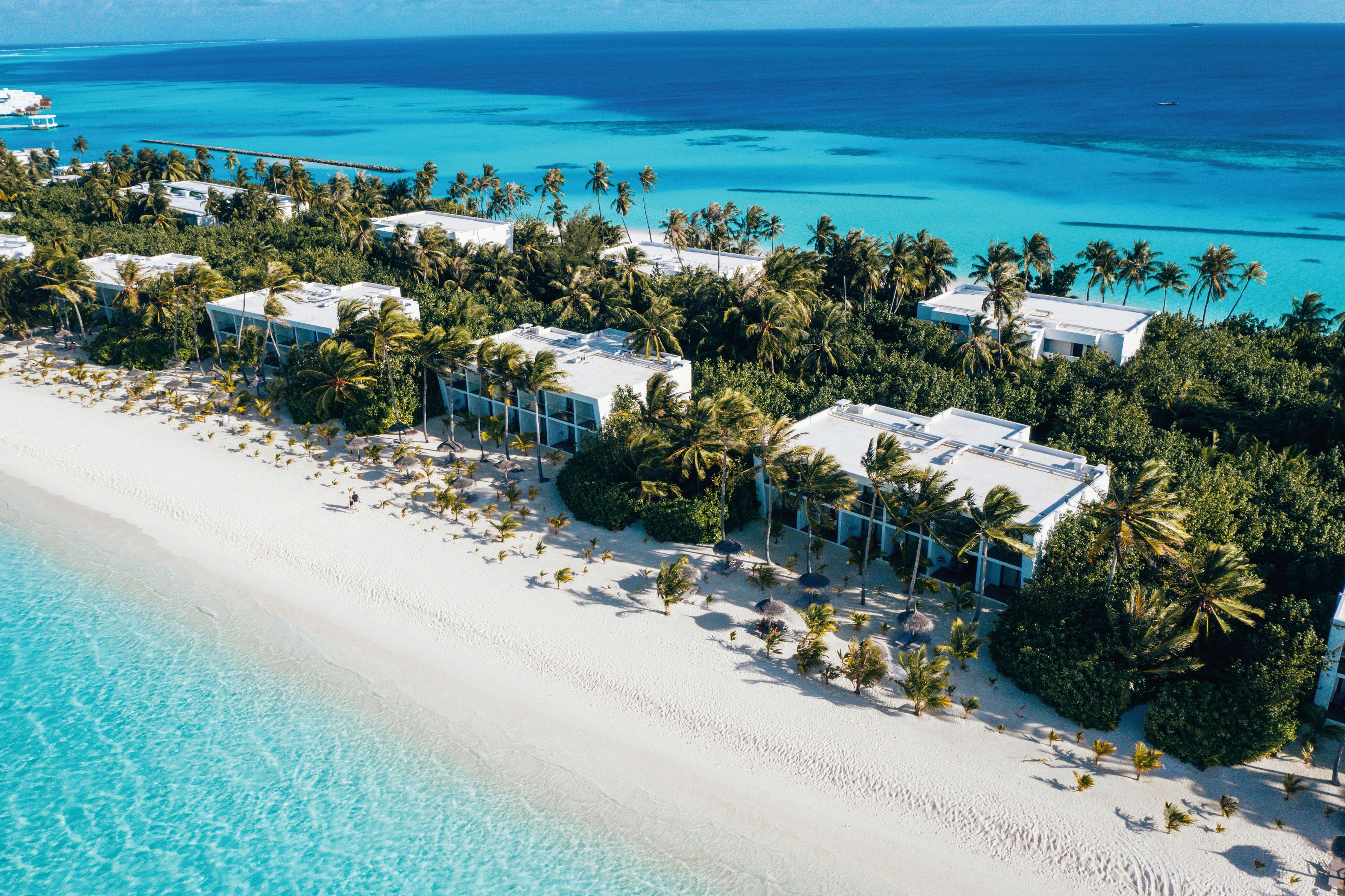 An aerial view of a beachfront resort with multiple villas, palm trees, and clear turquoise waters.