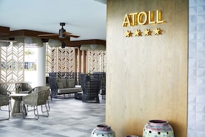 A modern indoor area with a wooden wall displaying the name "ATOLL" and five stars. There are wicker chairs and a table, and a ceiling fan is visible.