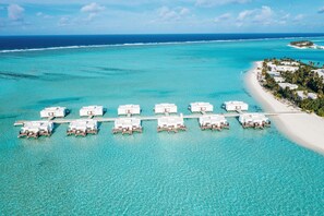 Vista aérea de un resort con bungalós sobre el agua alineados, conectados por un muelle, y una playa con palmeras.