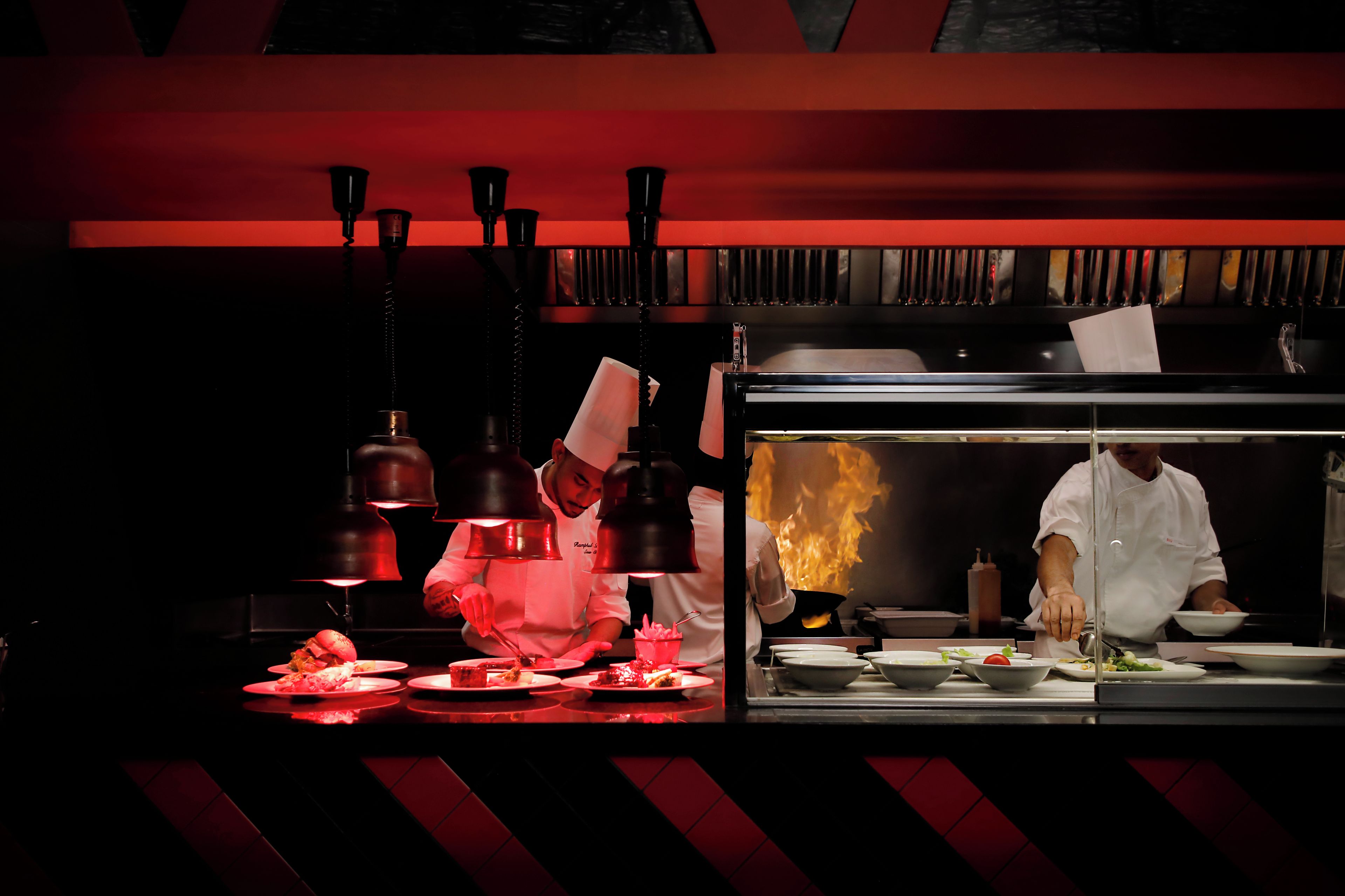 A chef preparing food in a kitchen with a red-lit background and a visible flame.