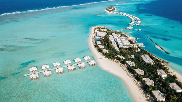 An aerial view of a resort island with overwater bungalows, a beach, and clear turquoise waters.