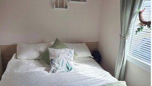 Uma cama bem arrumada com travesseiros e um cobertor, uma mesa de cabeceira, uma luminária na parede e uma janela com cortinas.
