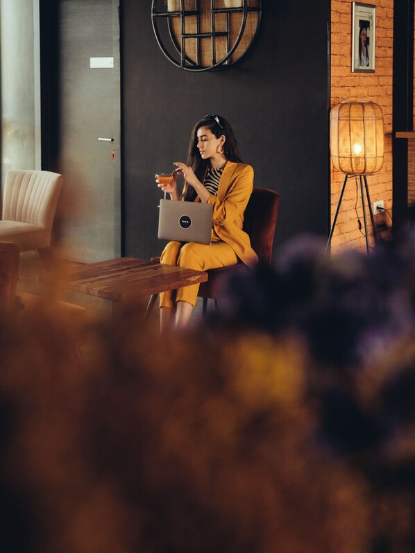 A person seated at a table with a laptop, a lamp, and a chair in a room with a brick wall and a round decorative item on the wall.