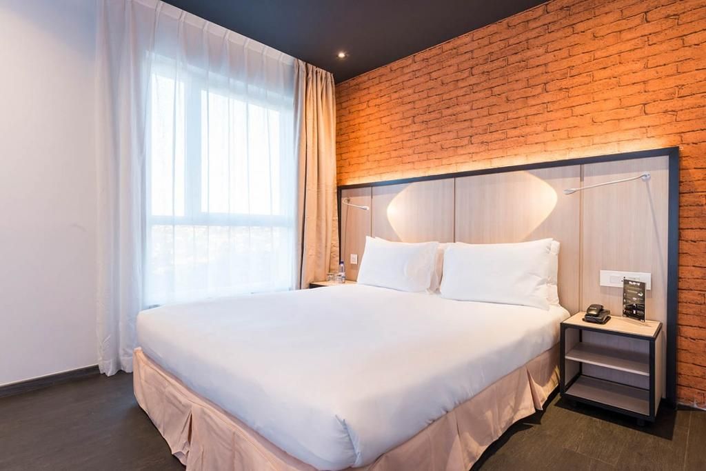 A modern hotel room with a large bed, a brick accent wall, a nightstand with a lamp, and a window with sheer curtains.