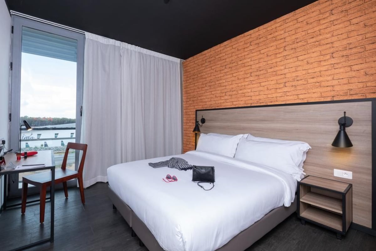 Quarto de hotel com cama grande, escrivaninha e cadeira. Há uma janela com cortinas, uma parede de tijolos e vista para um corpo de água.