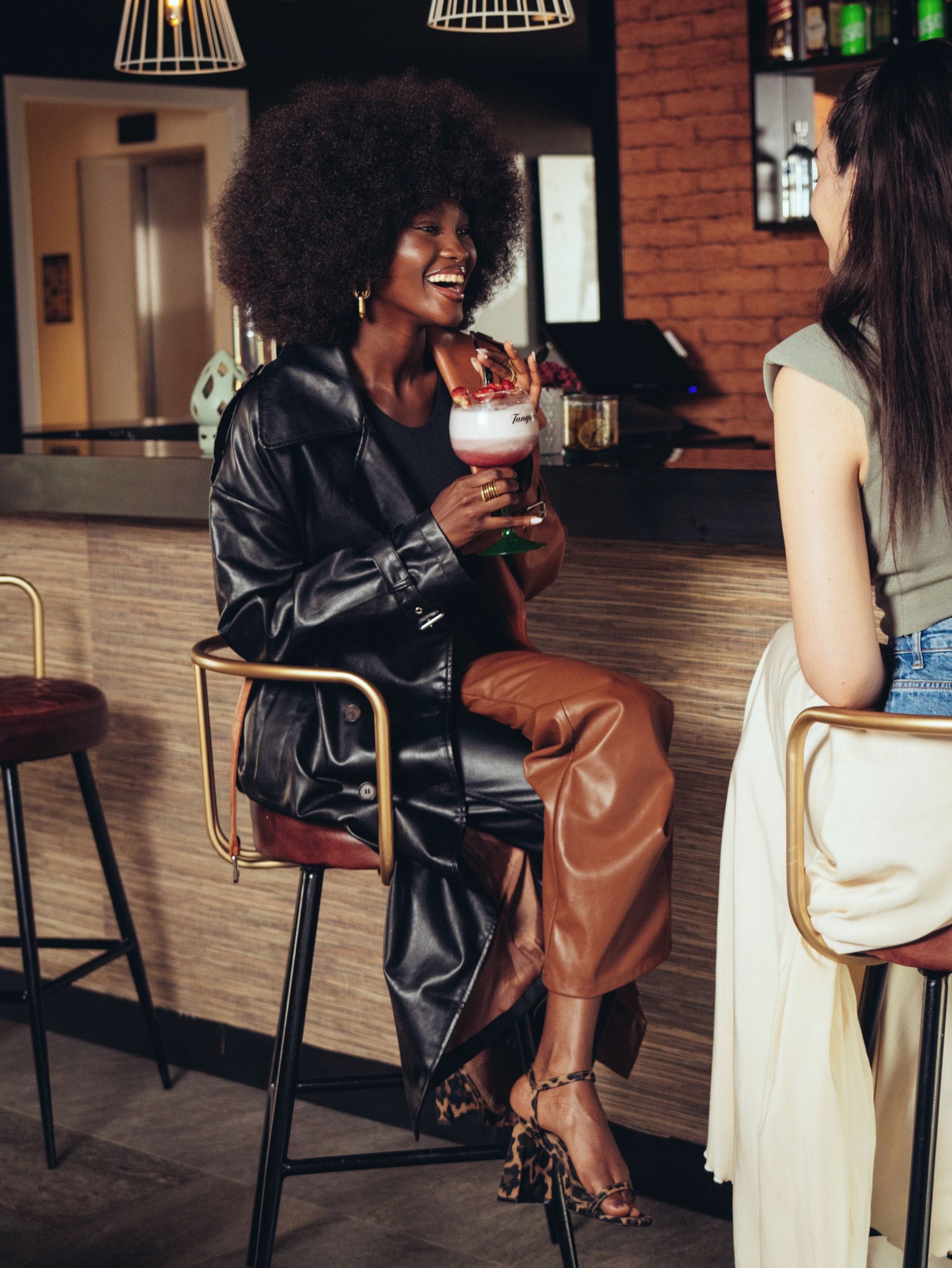 Une personne assise au bar, un verre à la main, une autre personne assise en face, et un espace bar avec des tabourets et un comptoir.