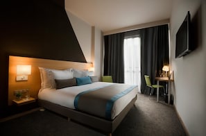 Quarto de hotel com cama, mesas de cabeceira, escrivaninha, cadeira, televisão e janela com cortinas.