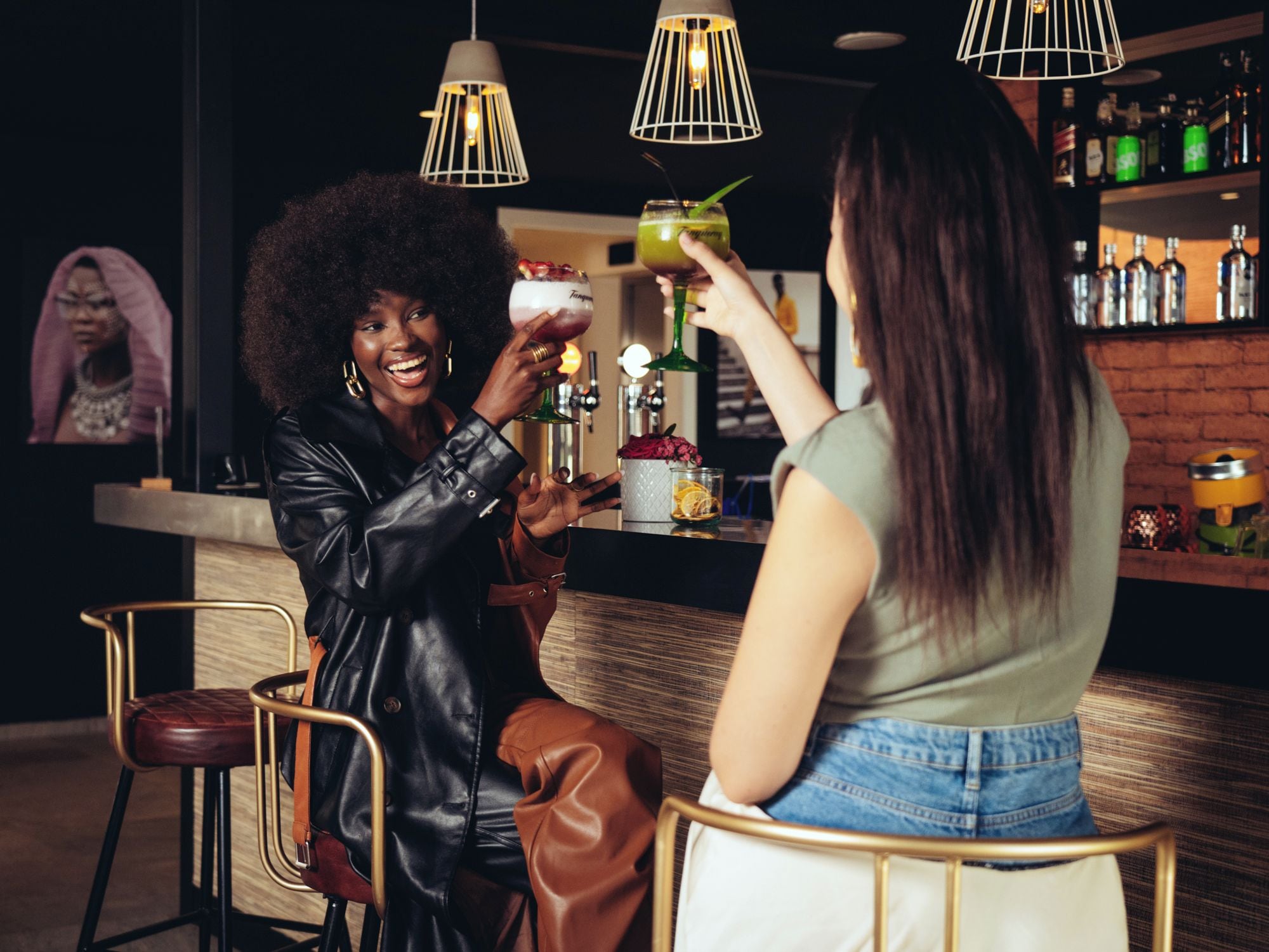 Zwei Personen sitzen an der Bar, jede mit einem Getränk in der Hand. Eine Theke mit Flaschen und eine Hängelampe sind ebenfalls zu sehen.