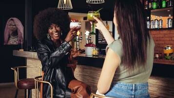 Zwei Personen sitzen an der Bar, jede mit einem Getränk in der Hand. Eine Theke mit Flaschen und eine Hängelampe sind ebenfalls zu sehen.