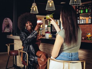 Dos personas sentadas en un bar, cada una con una bebida en la mano, una barra con botellas y una lámpara colgante arriba.