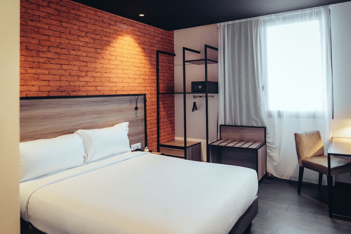 Quarto de hotel com parede de tijolos, uma cama, uma escrivaninha e uma cadeira.