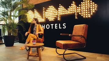 Uma pessoa sentada em uma cadeira de madeira com almofada, em frente a uma parede com a placa 'HOTELS' e uma luminária decorativa.